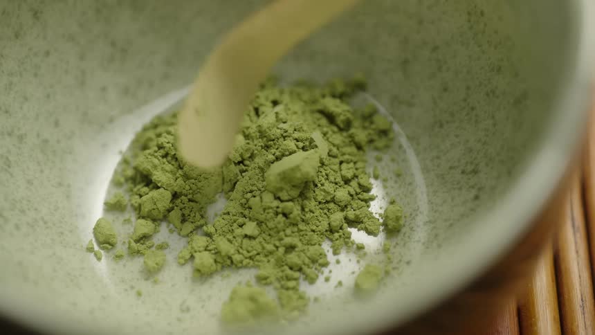 In slow motion, vibrant green matcha powder is carefully scooped with a bamboo utensil into a light green ceramic bowl