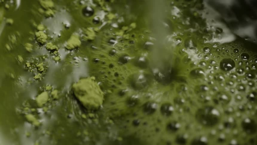 Hot water is poured over green matcha powder in a close-up, slow-motion shot, creating a bubbly tea