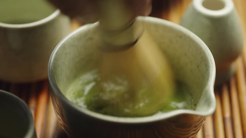 A hand vigorously whisks vibrant green matcha tea in a traditional ceramic bowl with a bamboo chasen