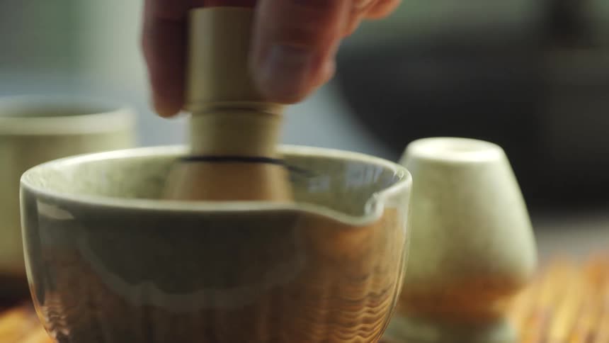 A close-up shot of a person preparing traditional Japanese matcha green tea by whisking it with a bamboo chasen in a ceramic bowl