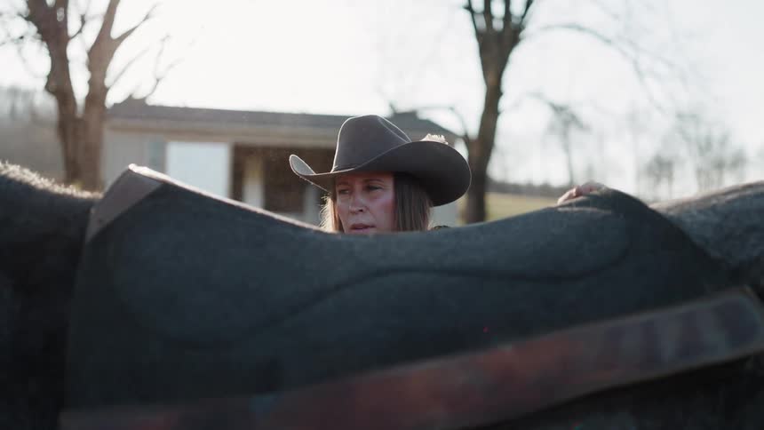 A focused cowgirl carefully places a saddle pad on her horse's back in slow motion on a dusty ranch