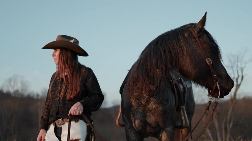 A cowgirl in full western attire stands peacefully with her saddled horse in a field during a beautiful golden hour