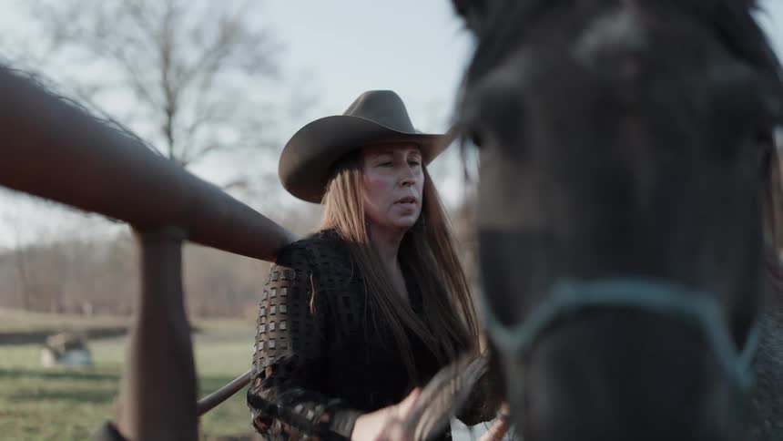 A woman in a cowboy hat stands by a metal fence and carefully grooms her horse in a pasture