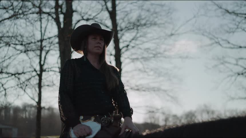 A stoic cowgirl sits on her horse in a rural setting, backlit by the late afternoon sun