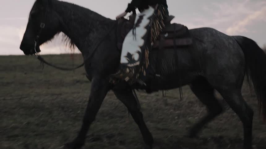 A cowgirl in a hat and chaps rides her dark horse through a field at dusk as the camera follows in slow motion