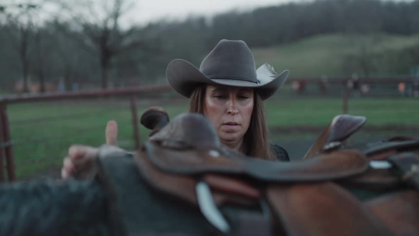 A determined cowgirl carefully removes the saddle from her horse in a paddock at dusk in slow motion