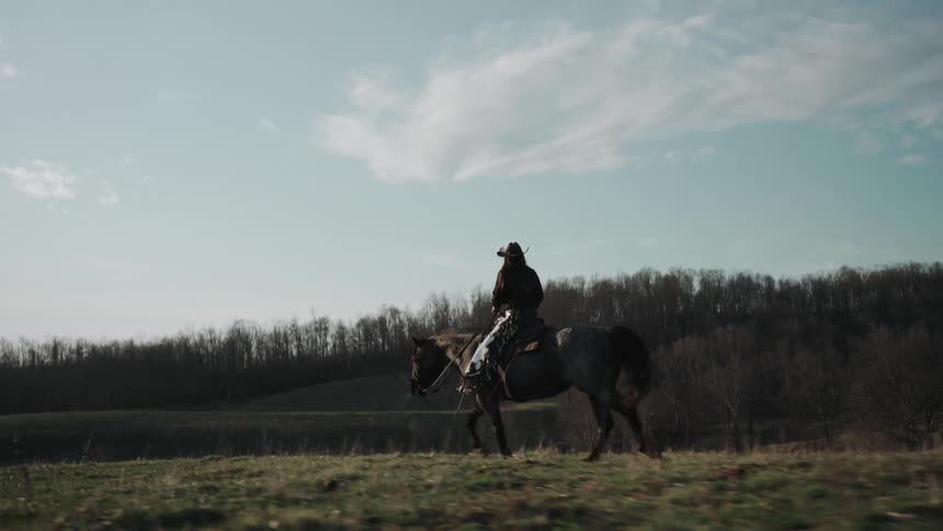 A lone cowgirl rides her horse across a grassy field in slow motion with rolling hills in the background
