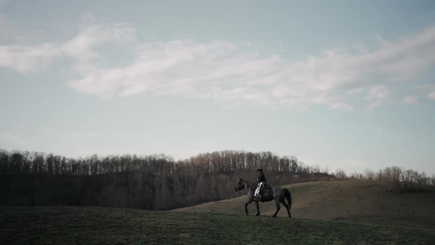 A lone rider in a cowboy hat slowly guides their horse across the crest of a grassy hill in the quiet countryside