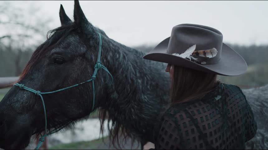 In slow motion, a woman wearing a feathered cowboy hat lovingly caresses her dark grey horse on an overcast day
