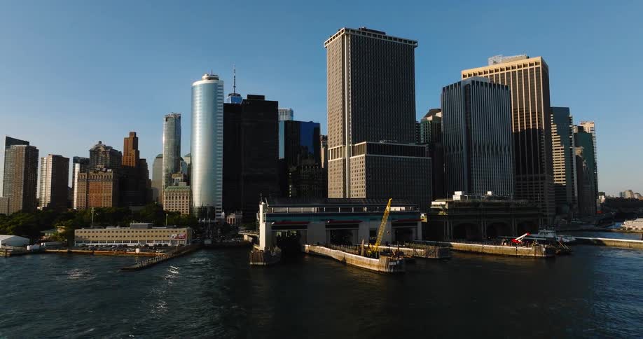 A majestic aerial view pulls back from the Whitehall Ferry Terminal, revealing the iconic Lower Manhattan skyline over the water on a bright, sunny day