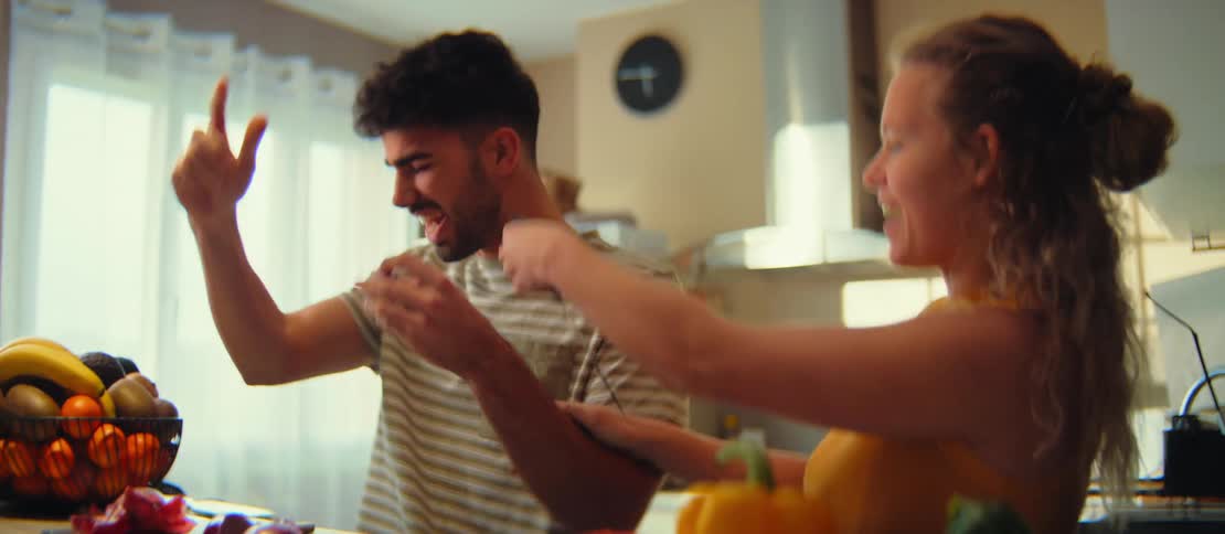 A joyful couple playfully sings and dances together in slow motion while preparing food in their sunlit kitchen