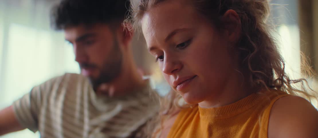 An intimate close-up of a young woman with curly hair focusing on a task, with a man blurred in the background