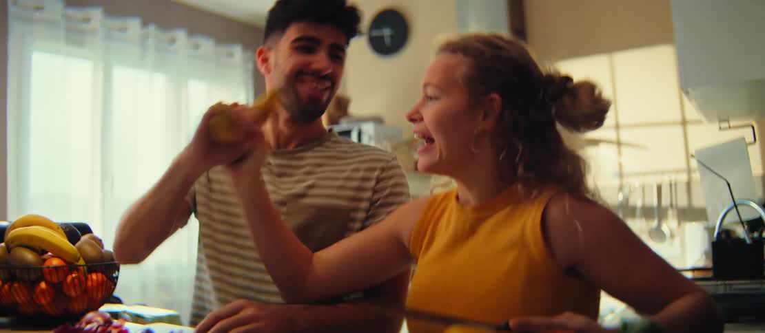A playful young couple laugh and sing into a banana while chopping vegetables together in their sunlit kitchen