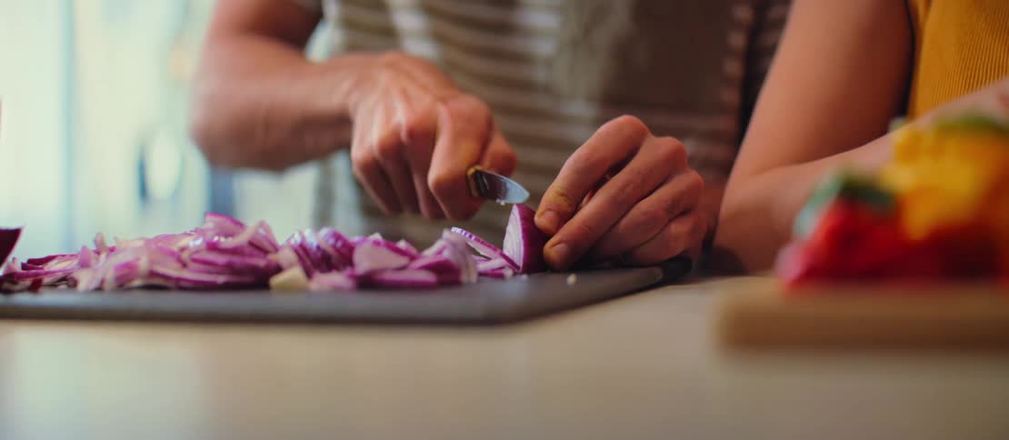 Slow motion close-up of a man's hands chopping a purple onion on a dark cutting board in a sunlit kitchen