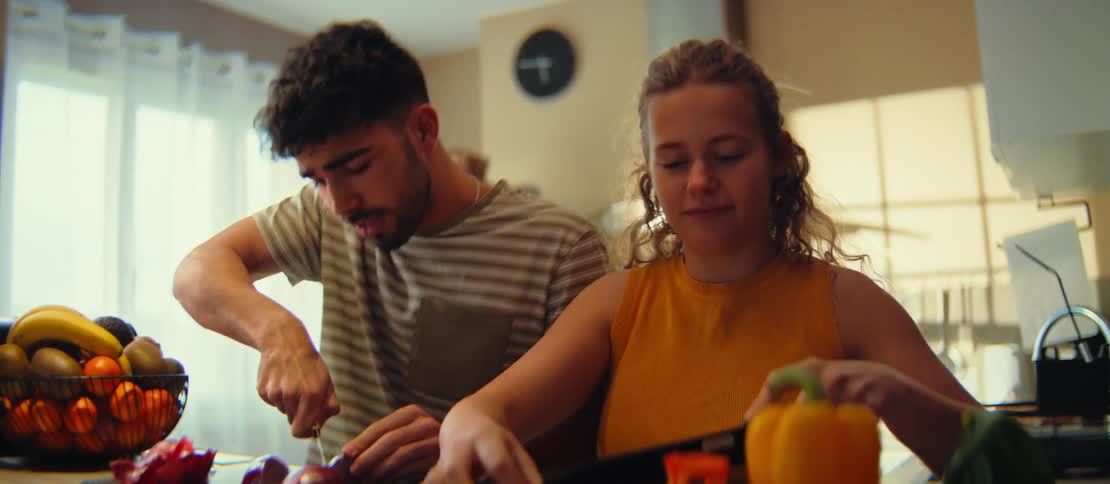 A smiling young couple enjoys cooking together, chopping fresh colorful vegetables on a cutting board in their bright kitchen