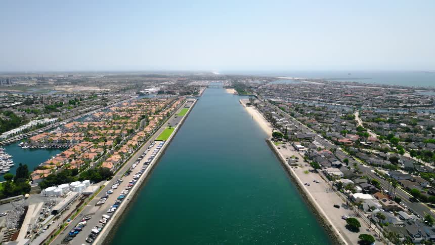 An expansive aerial view captures a long, straight waterway leading through a sunny coastal city with marinas and homes out to the vast ocean