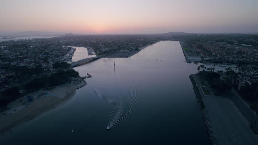 An aerial view captures a lone boat cruising through a tranquil harbor at dawn as the sun rises over the coastal city