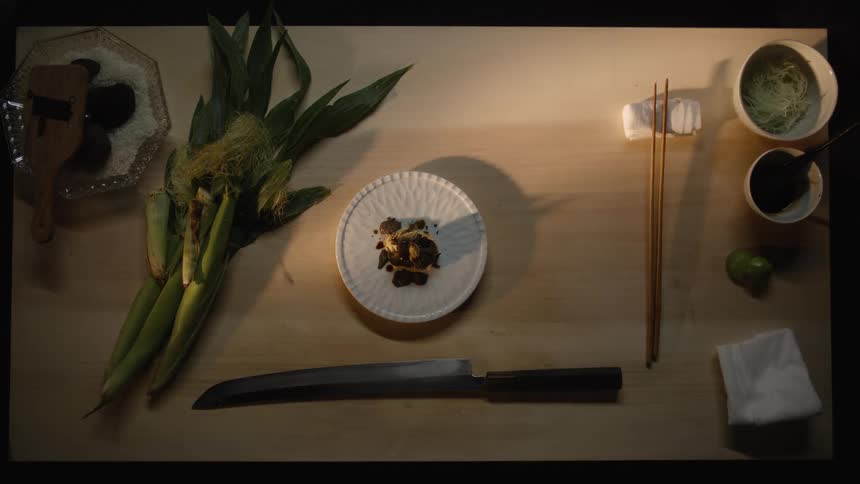 A dramatic top-down still life of fresh corn, black truffles, and a chef's knife arranged on a wooden surface