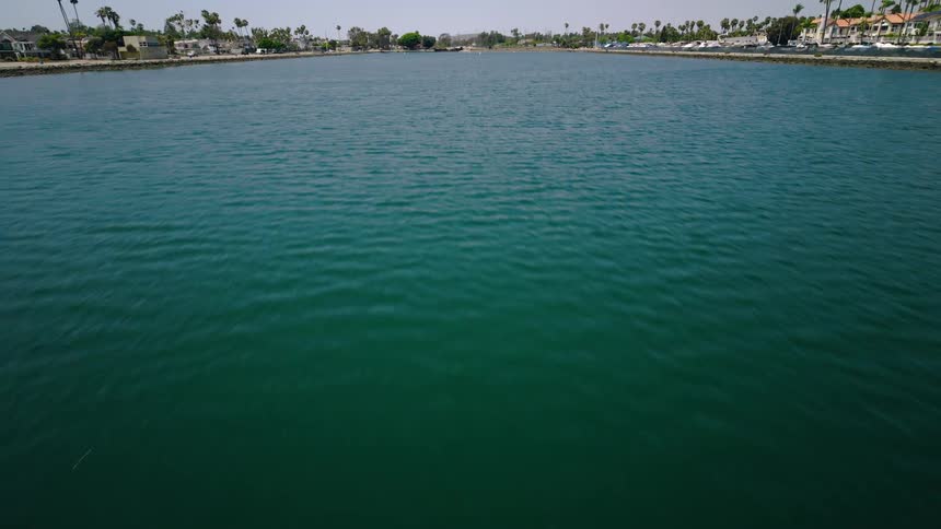 Aerial drone footage glides backward over the calm green water of a marina, slowly revealing the surrounding coastal community on a sunny day