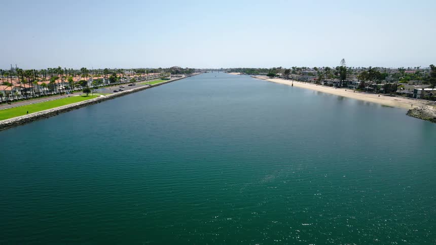 A calm, wide coastal channel flows between a sandy beach and a waterfront road on a bright, sunny day