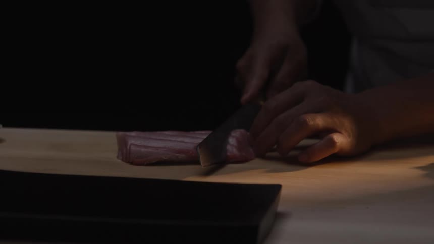 A sushi chef expertly slices a raw tuna fillet with a sharp knife on a wooden cutting board in dramatic slow motion
