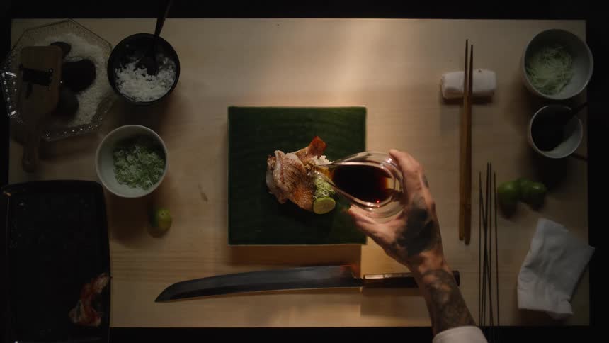 A tattooed chef's hands carefully pour soy sauce onto a beautifully plated piece of Japanese fish in this artistic, top-down cinemagraph