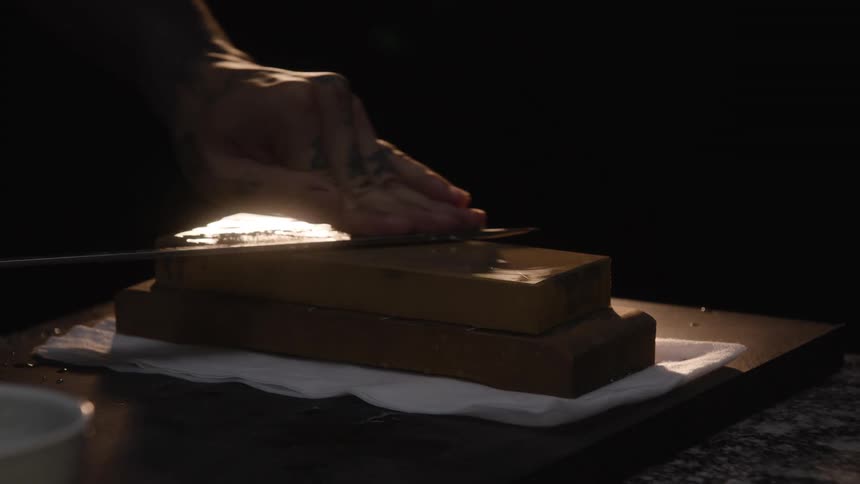 A person with tattooed hands carefully sharpens a long blade on a whetstone under a single spotlight in slow motion