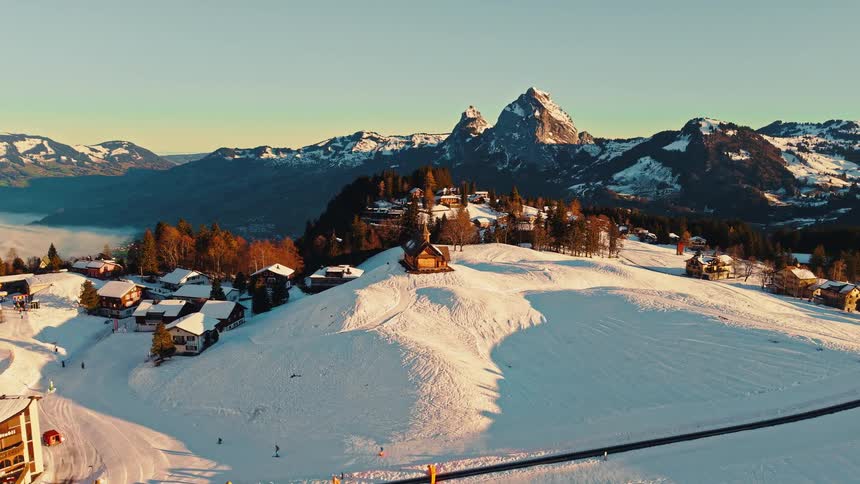 Stunning aerial drone footage pulls back over a snow-covered mountain village and ski resort in the Swiss Alps at sunrise