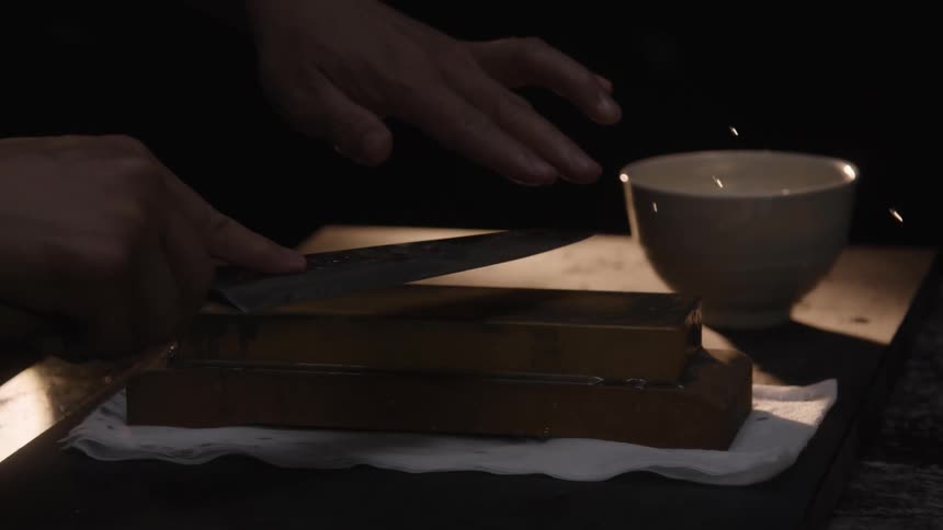 A chef pours water on a whetstone and meticulously sharpens a kitchen knife in dramatic slow motion