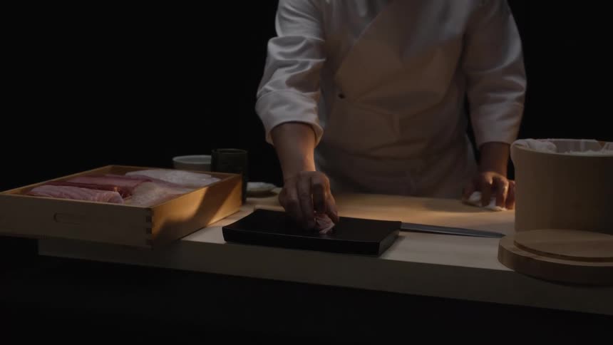 A skilled Japanese sushi chef meticulously prepares a piece of nigiri sushi in slow motion with a dark background