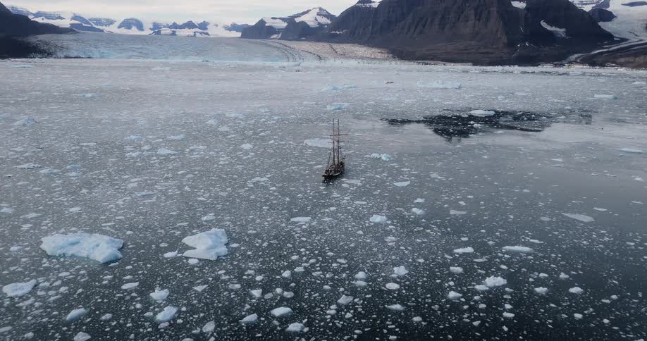 Aerial view of a historic sailing vessel surrounded by floating ice near a vast glacier and snowy mountains