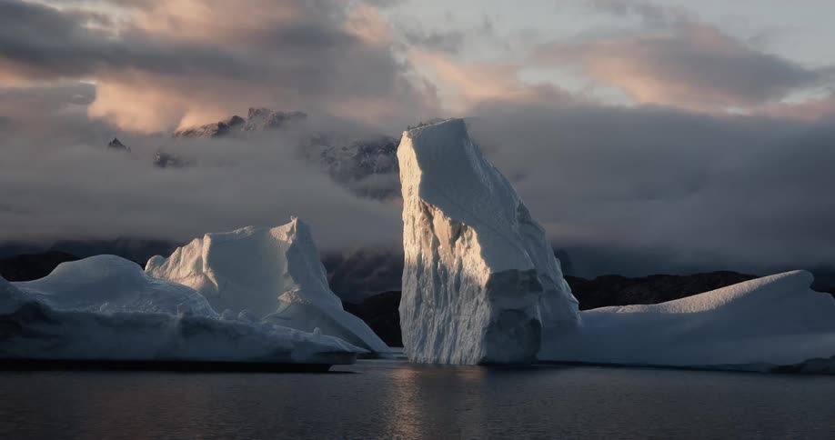 Majestic icebergs catch the golden light of sunrise in a calm arctic fjord with moody clouds and mountains