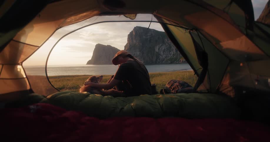 A young woman and her beloved dog share a quiet moment inside their tent, enjoying the sunset view over the ocean and cliffs