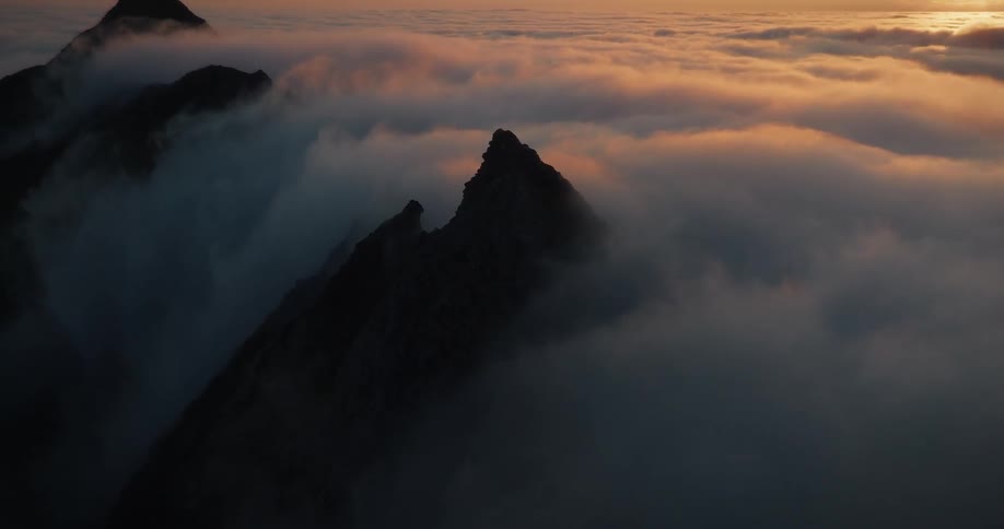 A stunning aerial shot slowly zooms in on a dramatic mountain peak rising above a sea of clouds during a golden sunset