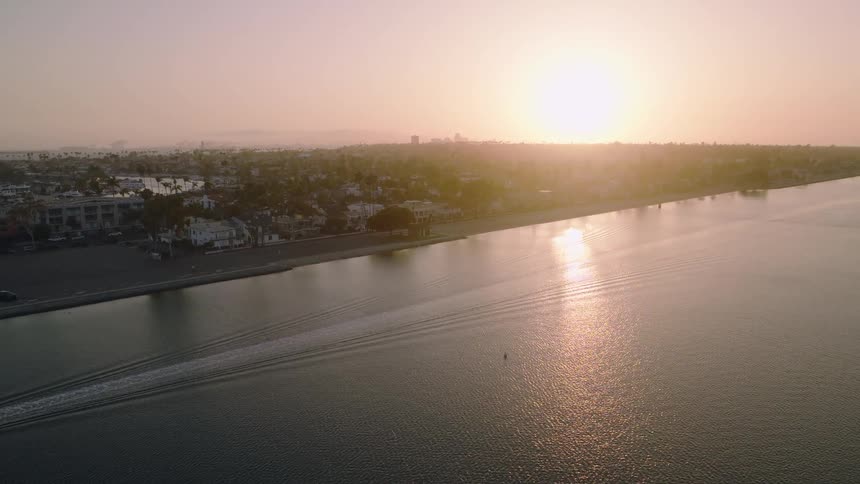 A serene aerial shot captures a motorboat gliding across the calm water of a bay during a hazy sunset