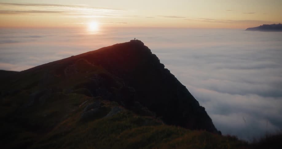 An epic wide shot shows a single person standing on a mountain summit above a sea of clouds during a golden sunrise