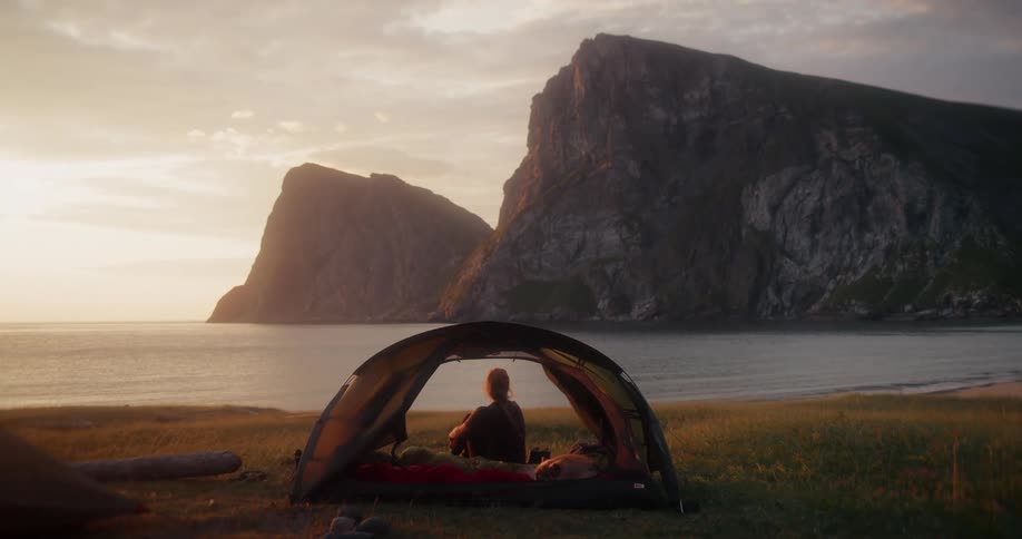 Lone camper sits peacefully in their tent enjoying the golden sunset over the tranquil ocean and majestic mountains