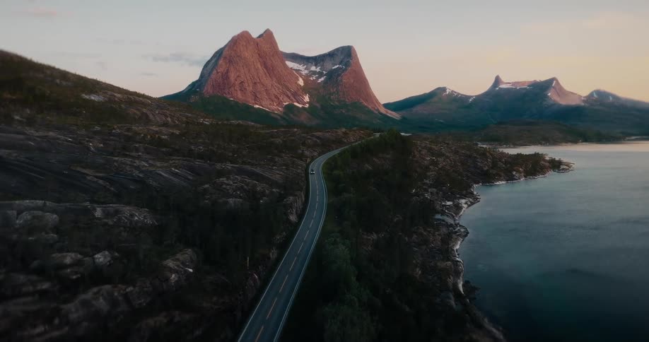 An aerial view shows a lone car driving on a scenic coastal road towards majestic, snow-capped mountains at sunset