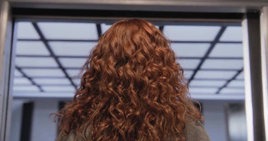 A back view of a woman with curly red hair as the modern elevator doors close in front of her