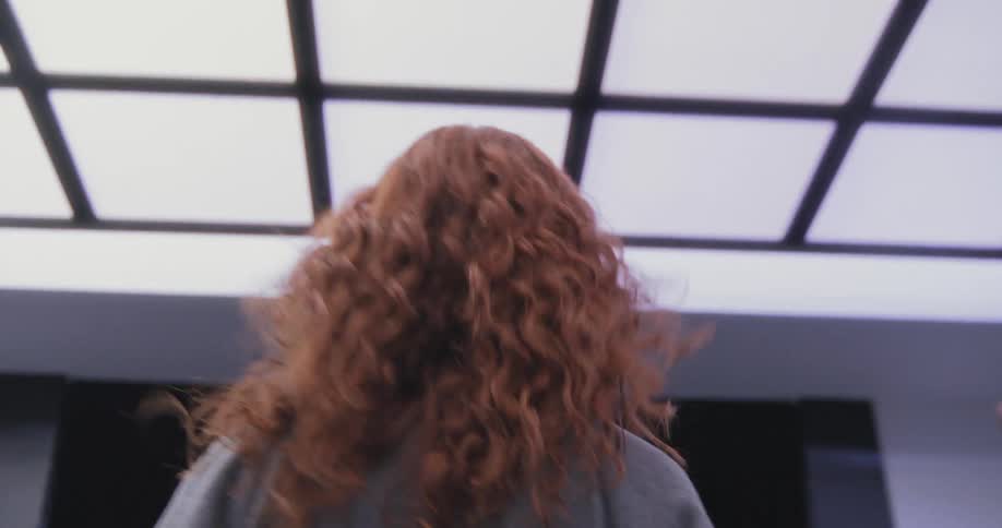 Low-angle tracking shot of a woman with curly red hair riding an escalator down from a brightly lit futuristic ceiling into darkness