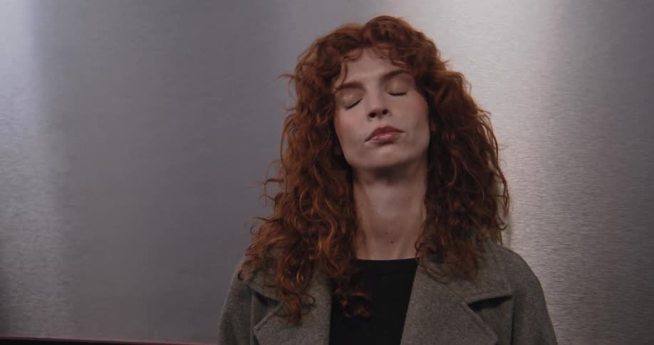 A weary woman with curly red hair leans against the wall in a mirrored room, appearing sad and introspective