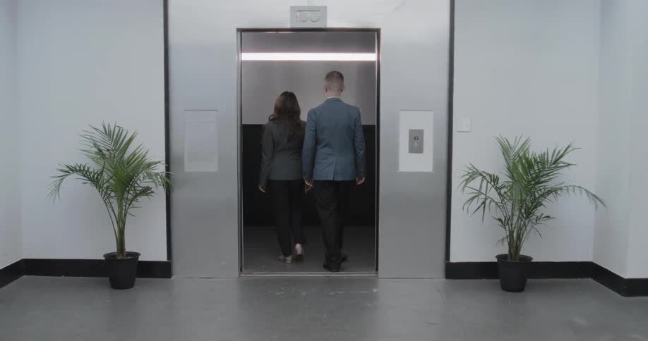A businessman and businesswoman walk from opposite directions to meet and enter an elevator in a modern office