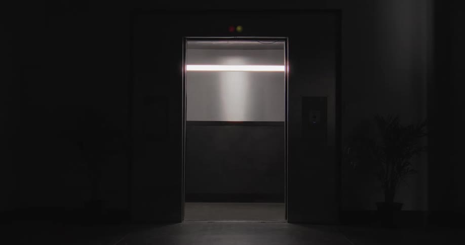 A modern elevator door slowly opens in a dark, empty hallway, revealing the lit interior