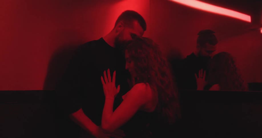 A passionate young couple kisses and embraces intimately while slow dancing in a dark room with sensual red lighting