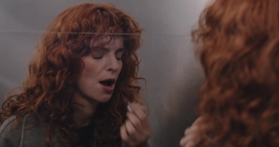 A stylish woman with vibrant red curly hair applies lipstick while looking at her reflection in an elevator mirror