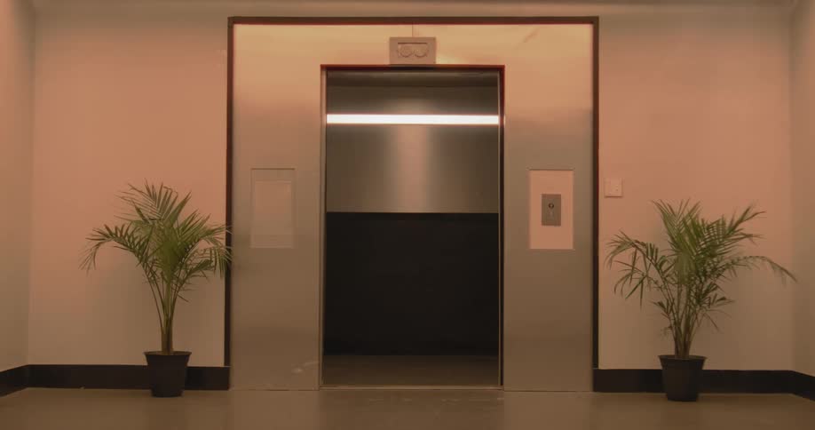 A static shot of a modern, stainless steel elevator as the doors slowly slide open in a warmly lit hallway