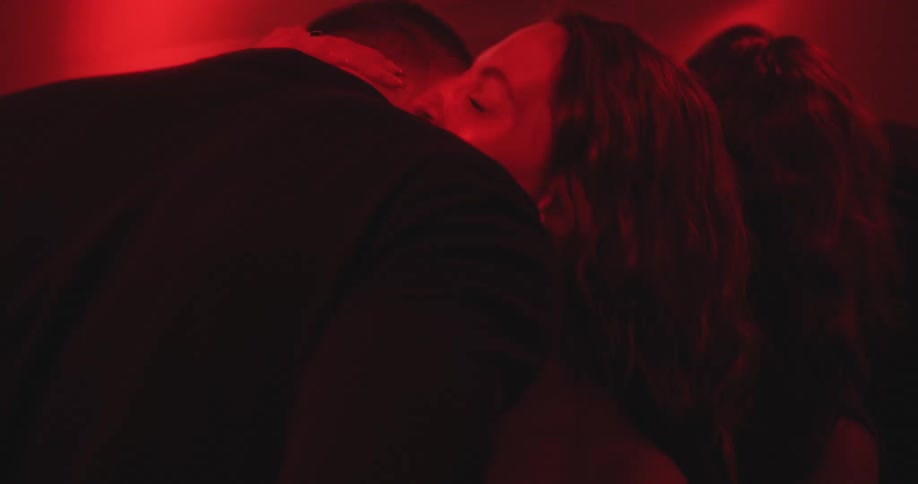 A passionate couple kisses and embraces in a dark, red-lit room, their reflections visible in the mirror