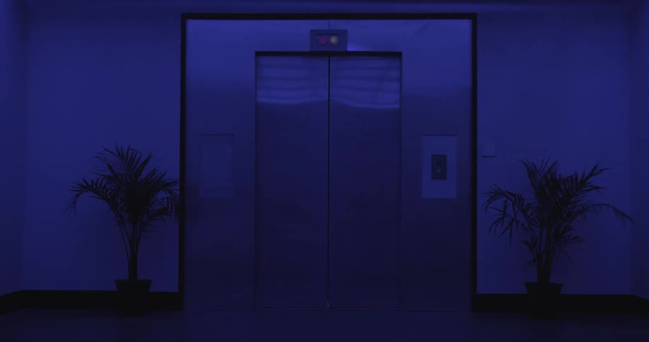 An empty modern elevator with stainless steel doors closes in a dark, blue-lit hallway