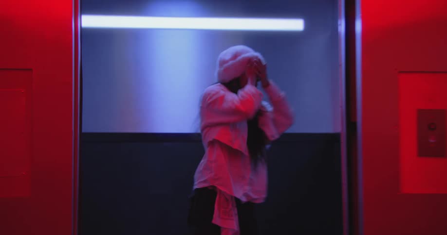 A man collapses in a red-lit room before the camera reveals a stylish woman dancing in an elevator