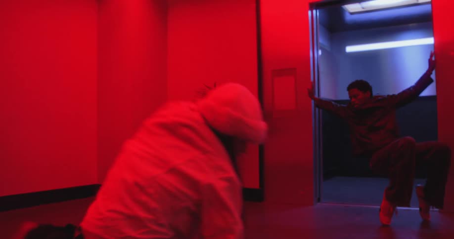 A stylish man and woman perform an energetic, expressive dance in slow motion within a modern hallway bathed in dramatic red light